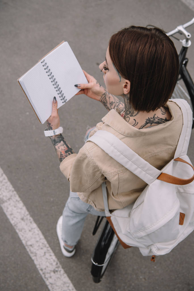 высокий угол обзора стильной татуированной женщины, держащей пустую тетрадь во время езды на велосипеде
  - Фото, изображение