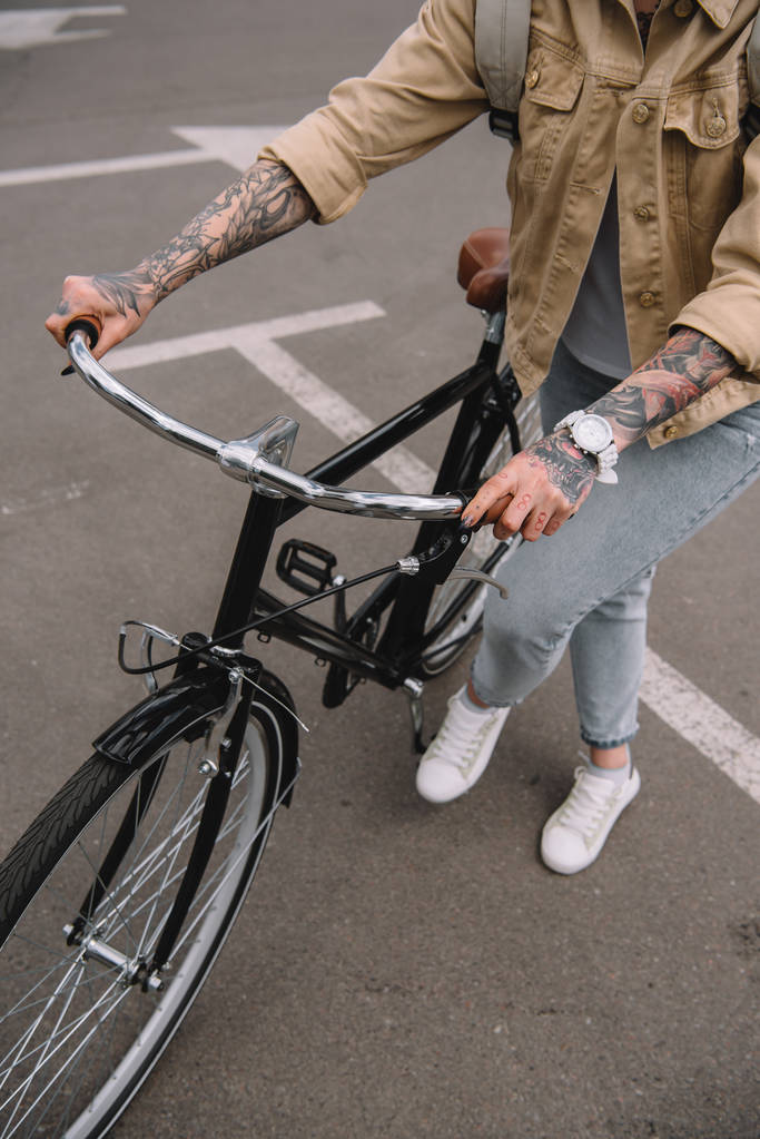 частичный вид женщины с татуировкой, стоящей с велосипедом на парковке
  - Фото, изображение