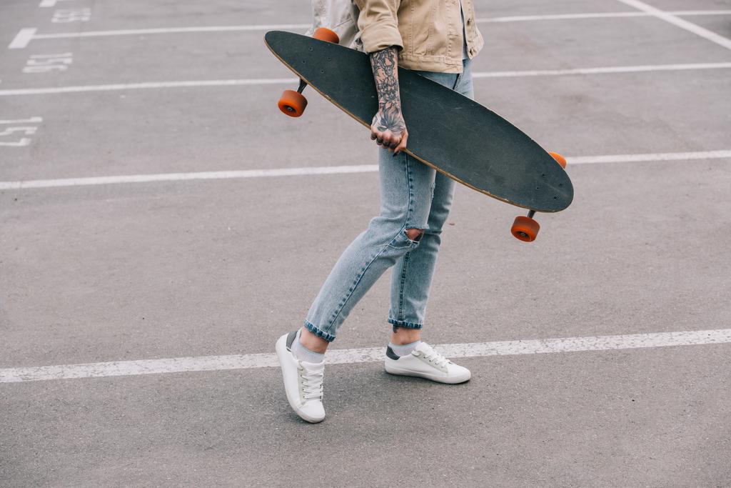 bijgesneden afbeelding van stijlvolle getatoeëerd vrouw houden longboard op parkeerplaats  - Foto, afbeelding