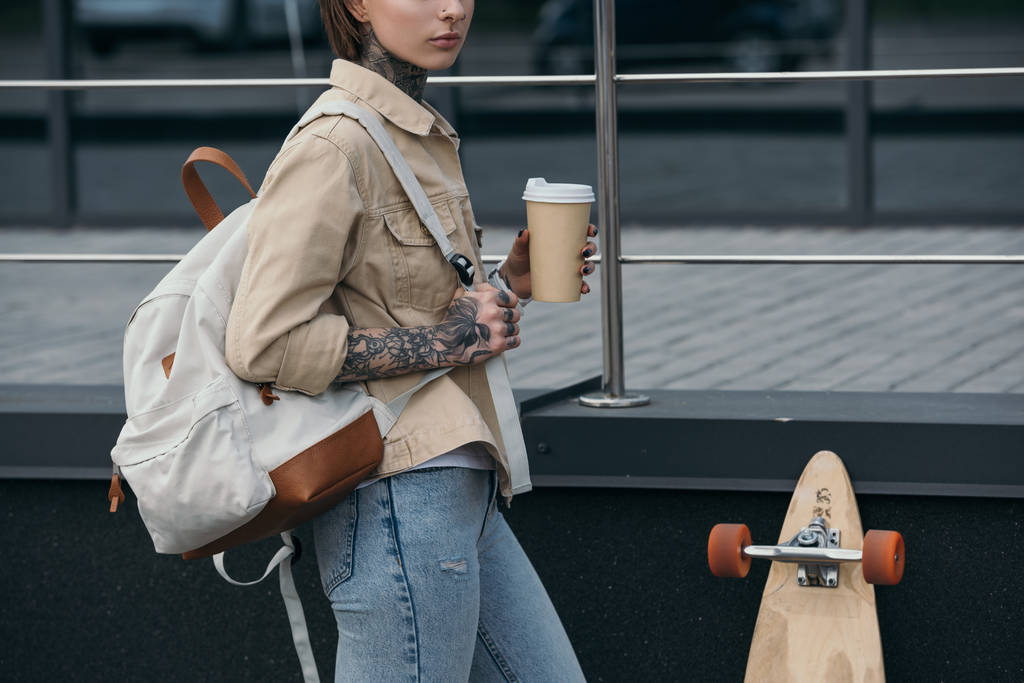 bijgesneden afbeelding van getatoeëerd vrouw met rugzak holding koffiekopje en permanent in de buurt van skateboard op de straat  - Foto, afbeelding