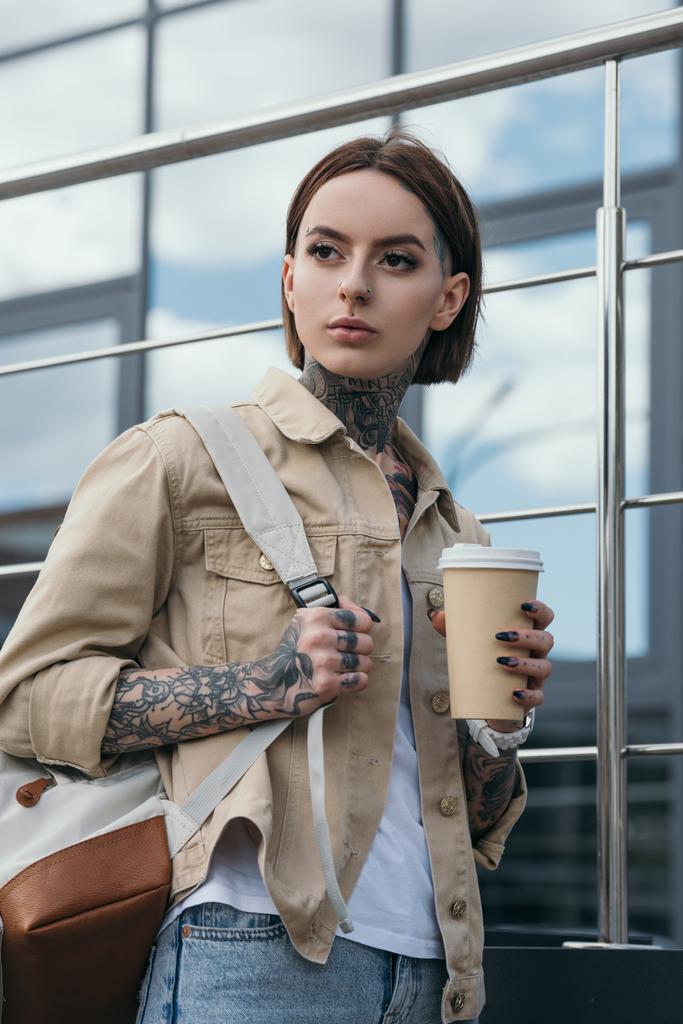 コーヒーの紙コップを保持しているバックパックとスタイリッシュな刺青女性のポートレート  - 写真・画像