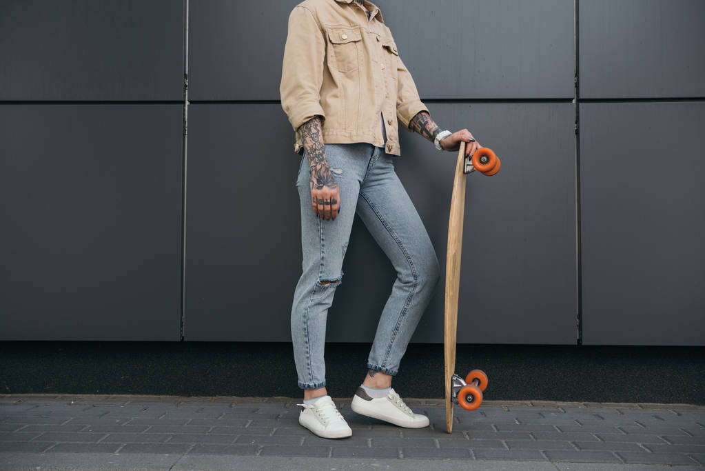 gedeeltelijke weergave van stijlvolle getatoeëerd vrouw stond met skateboard tegen zwarte muur  - Foto, afbeelding