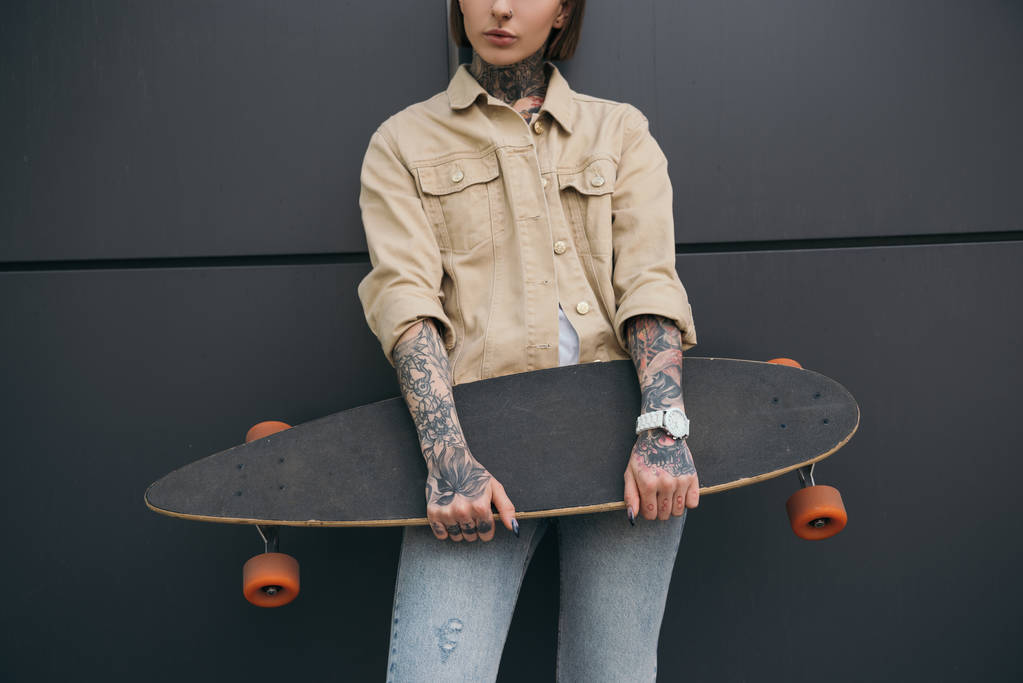 abgeschnittenes Bild tätowierter Frau, die Skateboard gegen schwarze Wand hält  - Foto, Bild