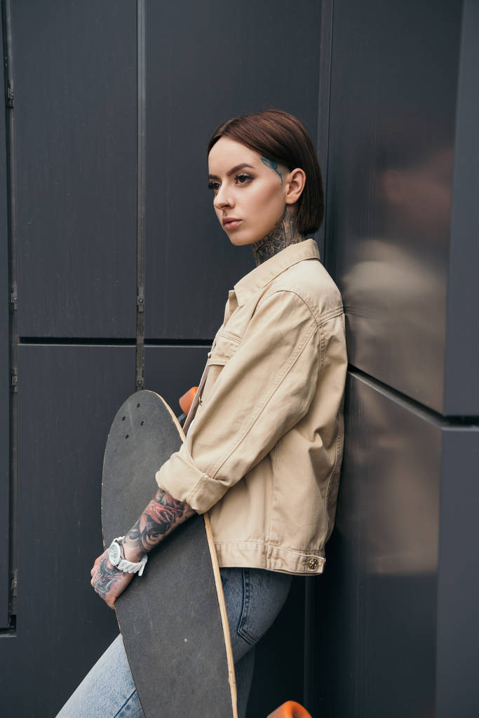 vista lateral de mujer tatuada sosteniendo monopatín contra pared negra
  - Foto, imagen