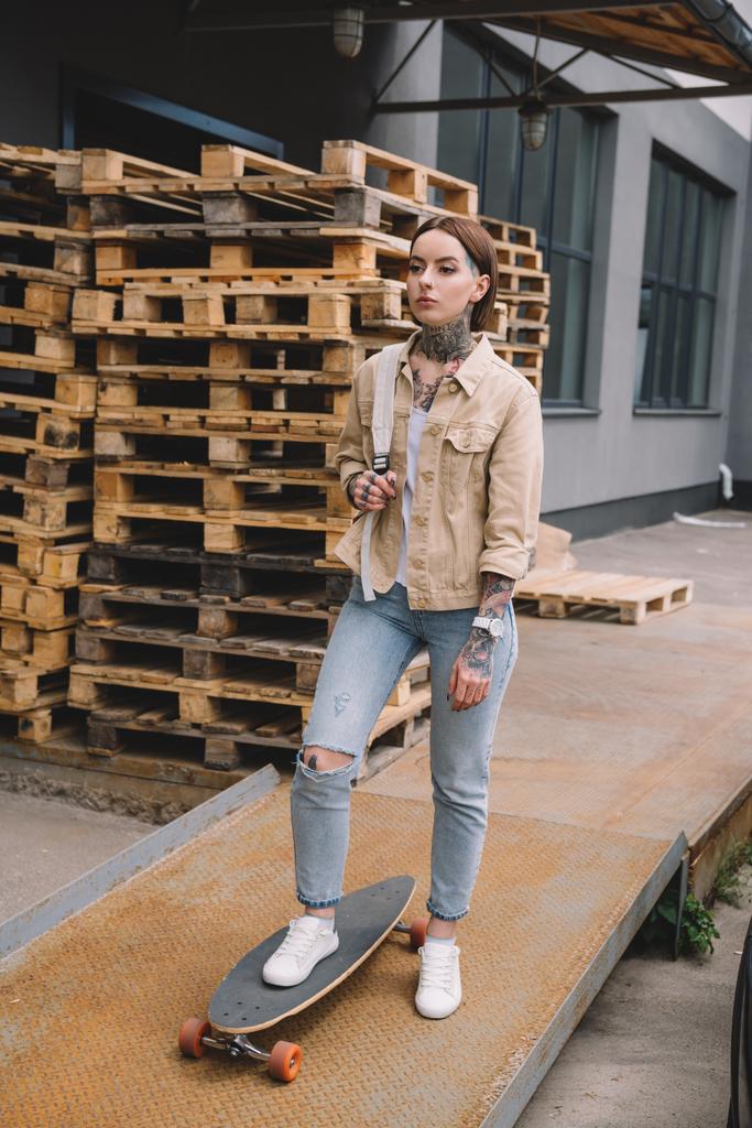 élégante femme tatouée debout avec planche à roulettes près de palettes en bois
  - Photo, image