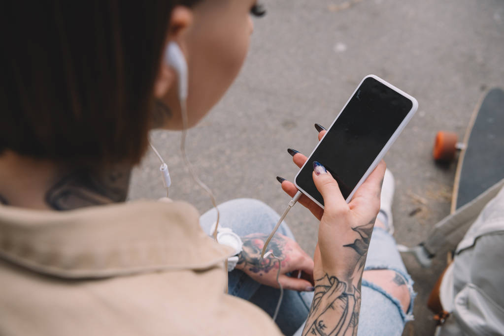 επιλεκτική εστίαση του τατουάζ γυναίκα στα ακουστικά, ακούγοντας μουσική και κρατώντας το smartphone με την κενή οθόνη - Φωτογραφία, εικόνα