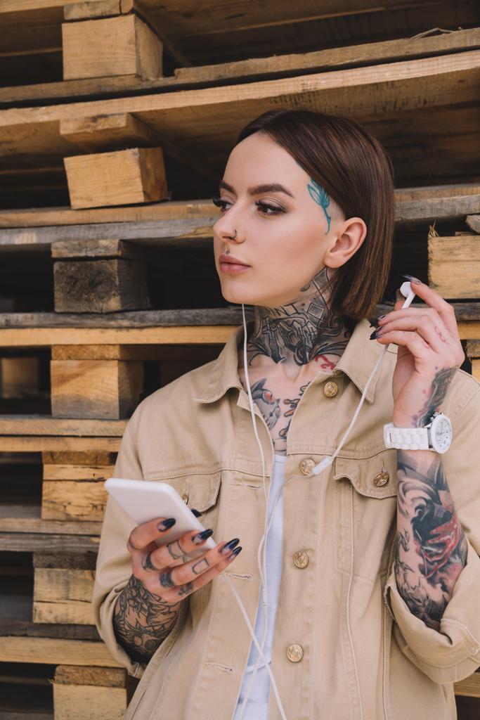 attractive tattooed woman in earphones listening music with smartphone  - Photo, Image