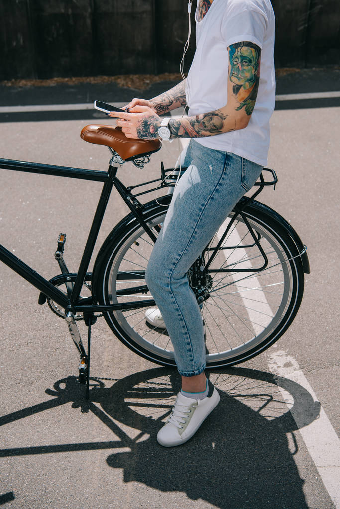 обрезанное изображение татуированной женщины в наушниках, слушающей музыку со смартфона, сидящей на велосипеде
  - Фото, изображение