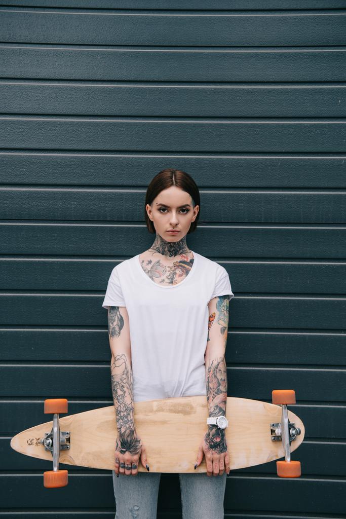 серьезная девушка с татуировками, держащая скейтборд и смотрящая в камеру на черную стену
 - Фото, изображение