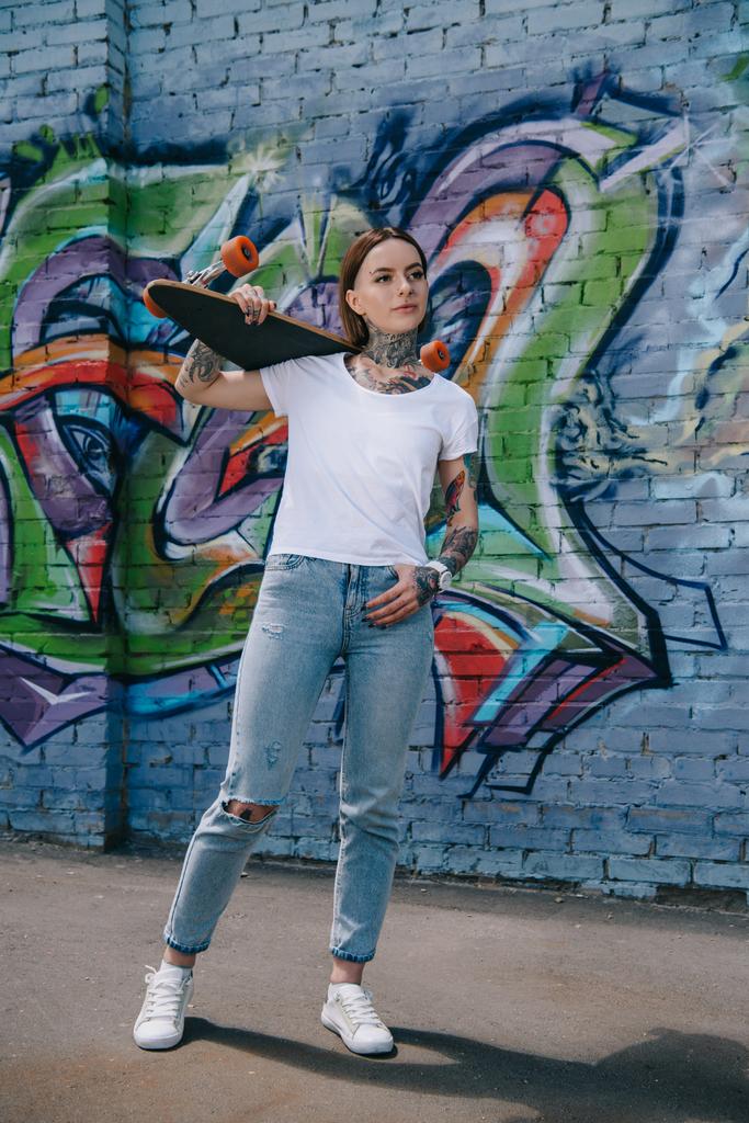 atraktivní dívka s tetováním držení skateboardu přes rameno zdi s graffiti - Fotografie, Obrázek