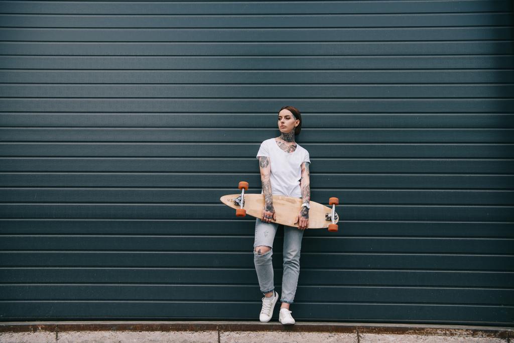 黒い壁にスケート ボードを持っての入れ墨を持つ若い女性の遠景  - 写真・画像