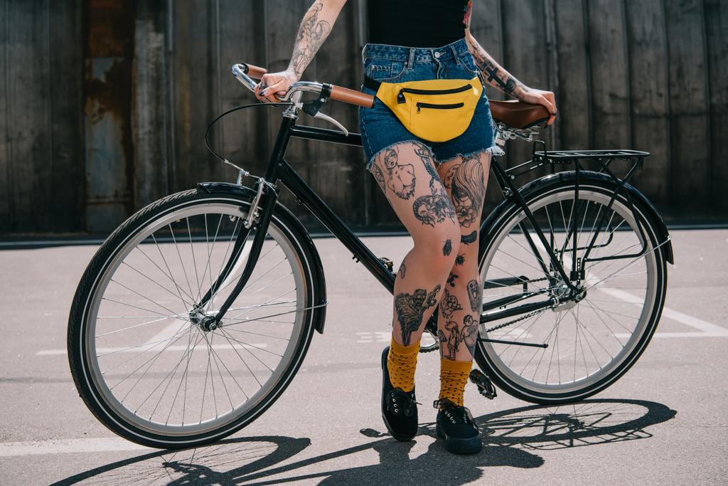 περικομμένη εικόνα κομψό τατουάζ κορίτσι με μέση bagstanding με ποδήλατο στο δρόμο - Φωτογραφία, εικόνα