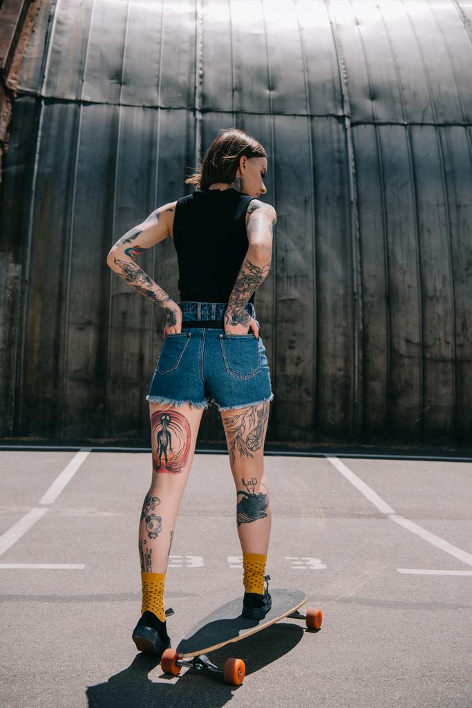 vista posteriore di elegante ragazza tatuata con le mani in tasche skateboard al parcheggio
  - Foto, immagini
