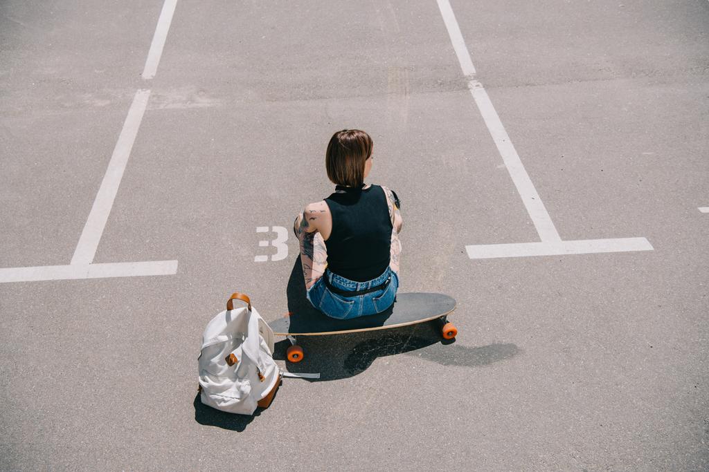 Achteraanzicht van stijlvolle getatoeëerd meisje zittend op het skateboard in de buurt van rugzak op parkeerplaats  - Foto, afbeelding