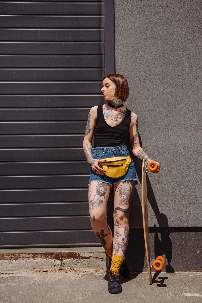 chica tatuada con estilo con bolsa de cintura que sostiene el monopatín contra la pared
  - Foto, Imagen