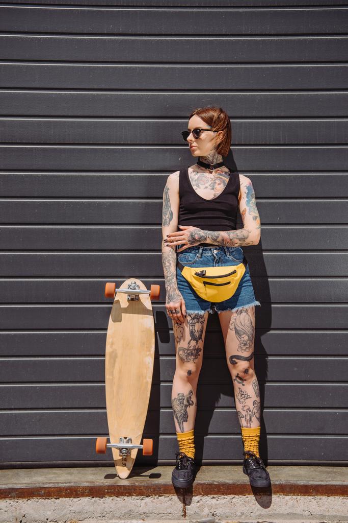 fille élégante avec des tatouages dans les lunettes de soleil regardant loin et debout près de planche à roulettes à la rue
 - Photo, image