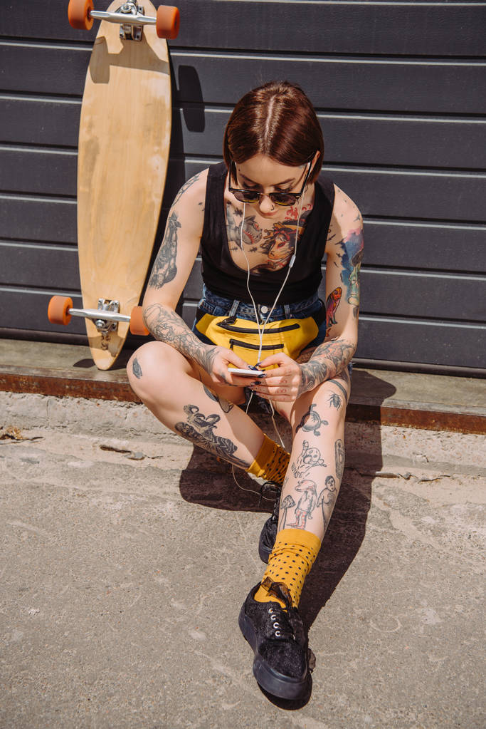 young tattooed woman in earphones listening music with smartphone near skateboard at street - Photo, Image