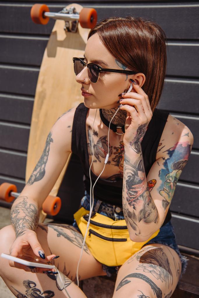 Tätowierte Frau mit Kopfhörer hört Musik mit Smartphone in der Nähe von Skateboard - Foto, Bild