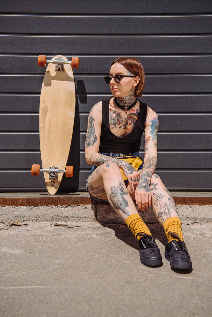 stijlvolle vrouw met tatoeages zitten in de buurt van skateboard op de straat - Foto, afbeelding