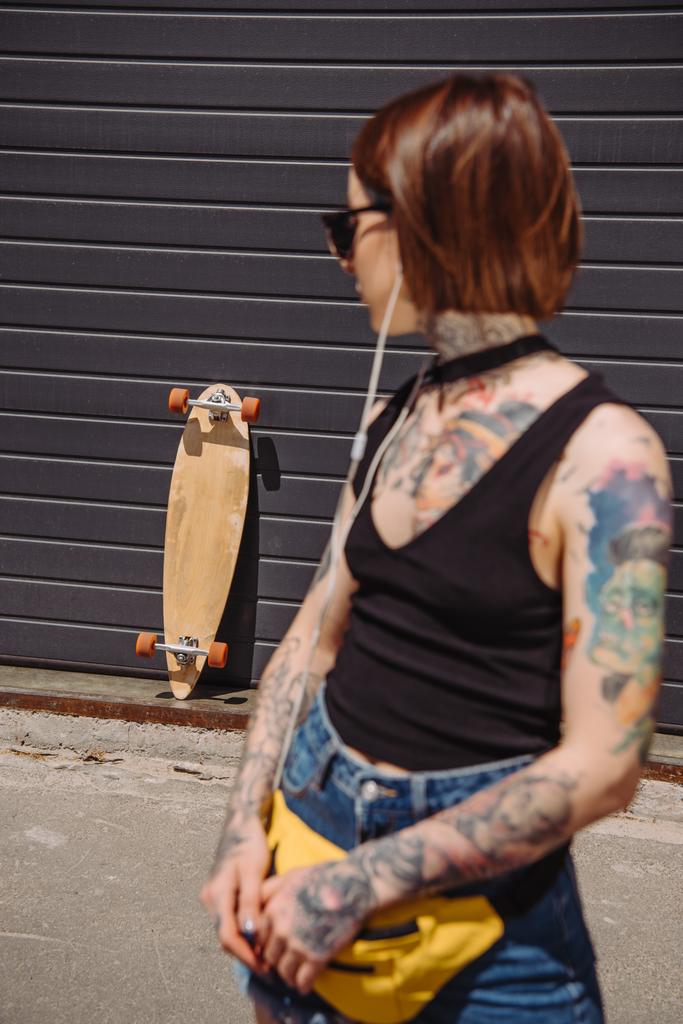 young tattooed woman listening music in earphones and looking at skateboard near wall  - Photo, Image