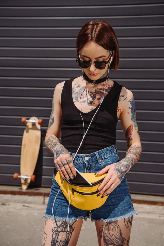 mujer tatuada con estilo escuchando música en los auriculares y poniendo teléfono inteligente en la bolsa de cintura
  - Foto, Imagen