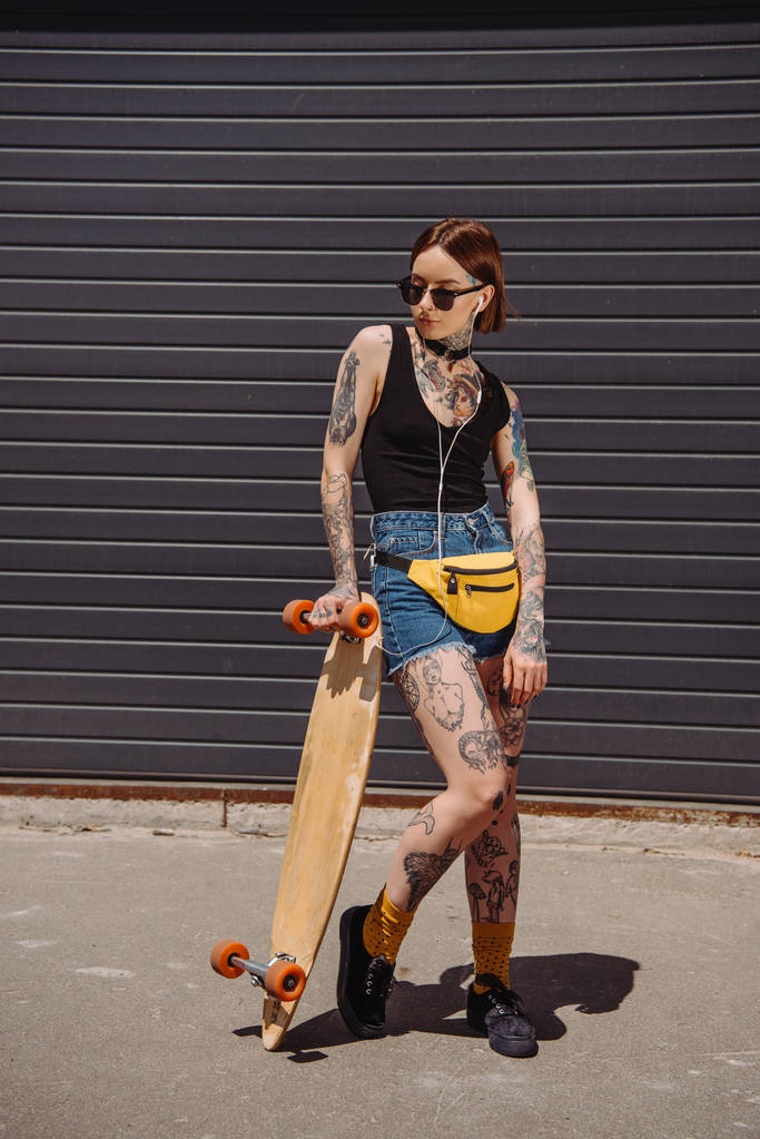 stylowej kobiety tatuażem na okulary przeciwsłoneczne, słuchanie muzyki za pomocą słuchawek i stojący w pobliżu deskorolka  - Zdjęcie, obraz