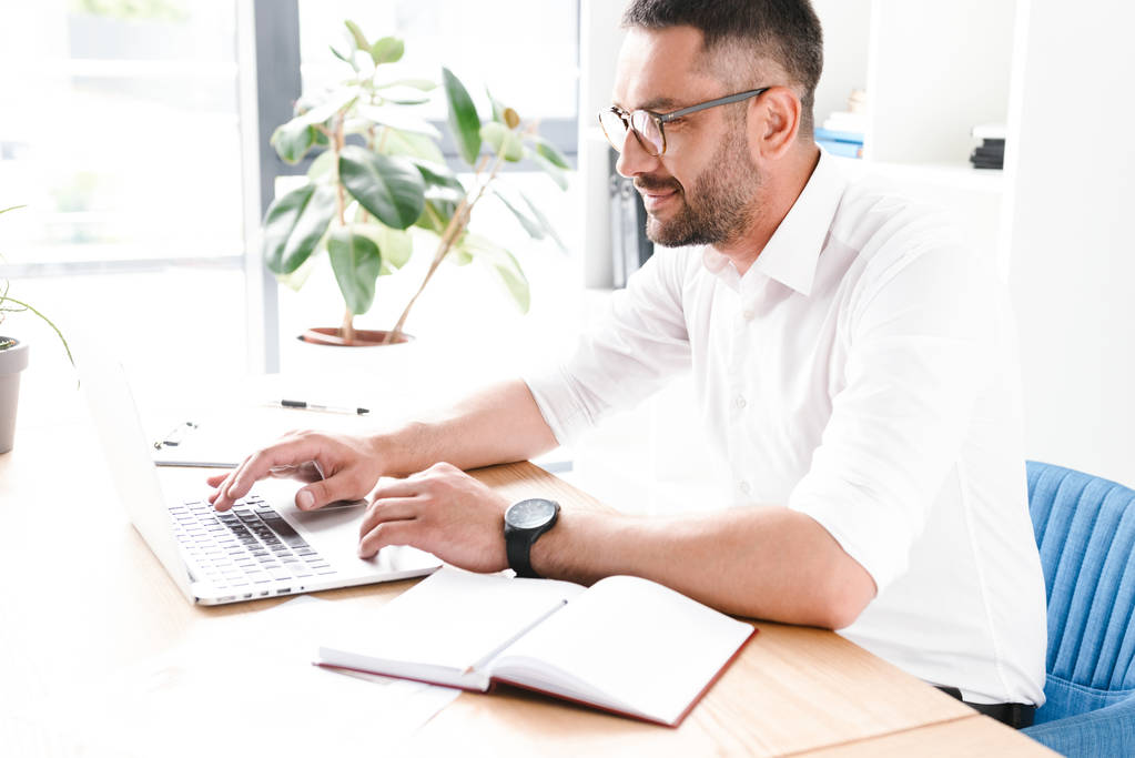 Kuva menestyneestä aikuisesta miehestä 30s valkoisessa paidassa istuu pöydässä ja käyttää kannettavaa tietokonetta työskennellessään valoisassa toimistohuoneessa
 - Valokuva, kuva