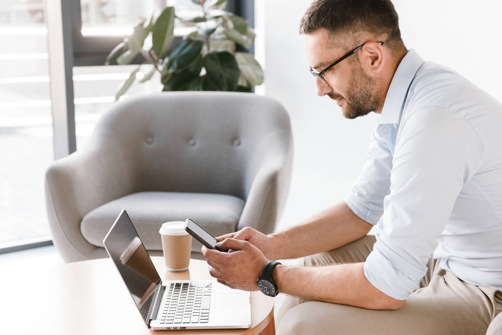 Horizontale foto van zakenman dragen van bril zitten in een leunstoel, en het gebruik van de smartphone tijdens het werken op de laptop in kantoor interieur - Foto, afbeelding