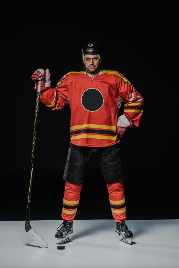 volle lengte weergave van professioneel ijshockeyspeler permanent met hand op de taille en camera kijken op zwart - Foto, afbeelding