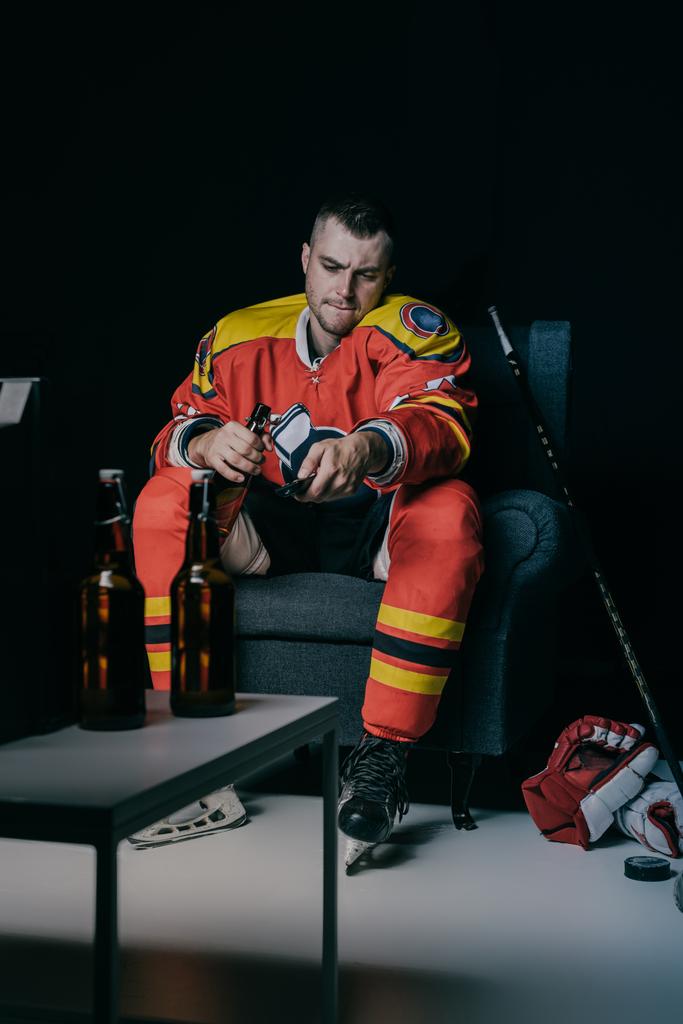 young hockey player using remote controller and drinking beer while sitting in armchair and watching tv on black - Photo, Image