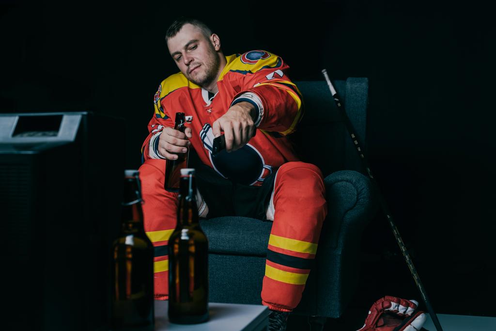 hockey player using remote controller and drinking beer while sitting and watching tv on black - Photo, Image