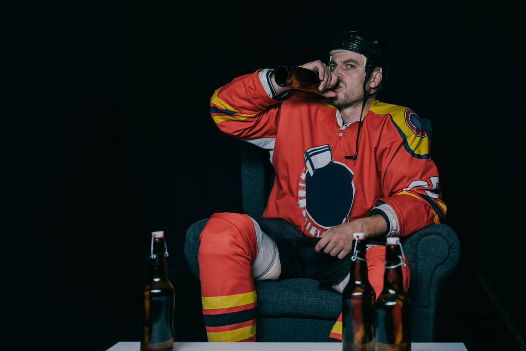 giovane giocatore di hockey bere birra mentre seduto in poltrona e guardare la tv su nero
 - Foto, immagini