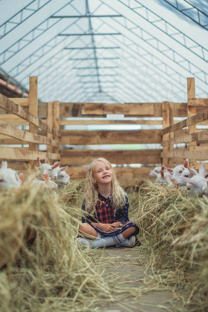 Lächelndes Kind sitzt auf dem Boden in der Nähe von Ziegen auf dem Bauernhof und schaut weg - Foto, Bild
