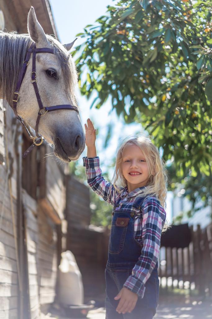 smiling kid touching white horse at farm and looking at camera - Photo, Image