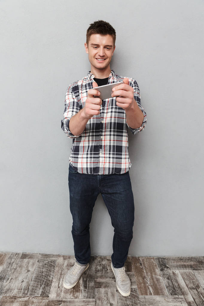 Täyspitkä muotokuva hymyilevä nuori mies pelaa pelejä matkapuhelimella harmaalla taustalla
 - Valokuva, kuva