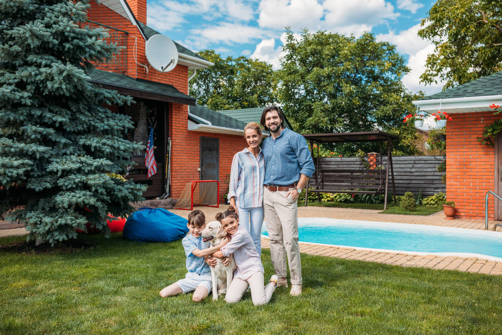χαρούμενη οικογένεια με σκυλί λαμπραντόρ βλέπουν φωτογραφική μηχανή ξοδεύοντας το χρόνο στην πίσω αυλή του σπιτιού χώρας θερινή ημέρα - Φωτογραφία, εικόνα