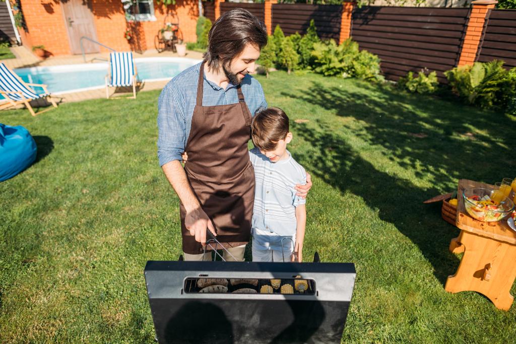 son looking at father cooking sausages and corn on grill on backyard - Photo, Image