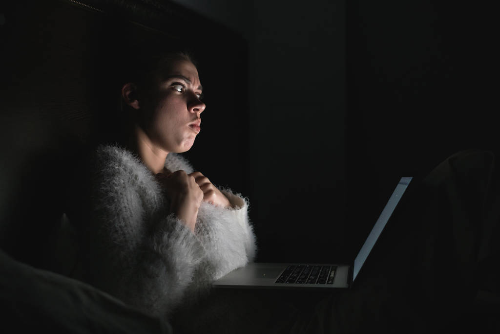 φοβισμένη νεαρή κοπέλα κάθεται στο κρεβάτι αργά τη νύχτα, βλέποντας ταινία τρόμου στο φορητό της, στο σκοτάδι - Φωτογραφία, εικόνα