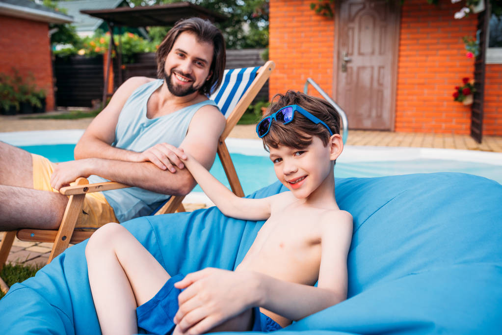uśmiechnięty ojciec i syn odpoczynku w pobliżu basen na podwórku na letni dzień - Zdjęcie, obraz