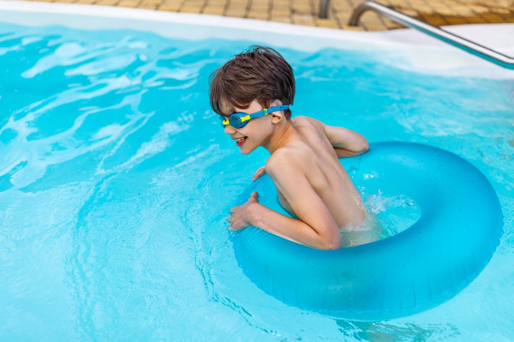 Zijaanzicht van gelukkige jongen in zwemmen bril met opblaasbare ring zwemmen in zwembad op zomerdag - Foto, afbeelding