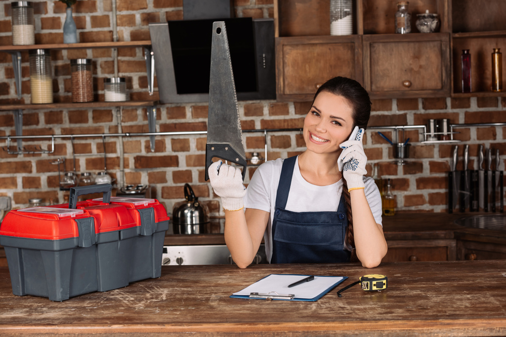 happy young repairwoman with saw taking by phone at kitchen - Photo, Image