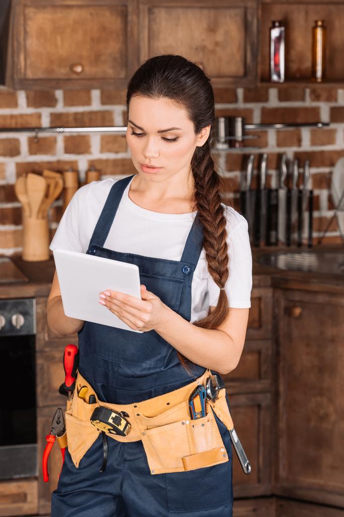 ernstige jonge repairwoman met toolbelt met behulp van Tablet PC op keuken - Foto, afbeelding