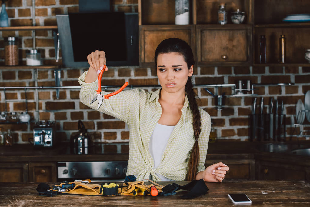 mujer joven confundida mirando alicates en la cocina
 - Foto, Imagen