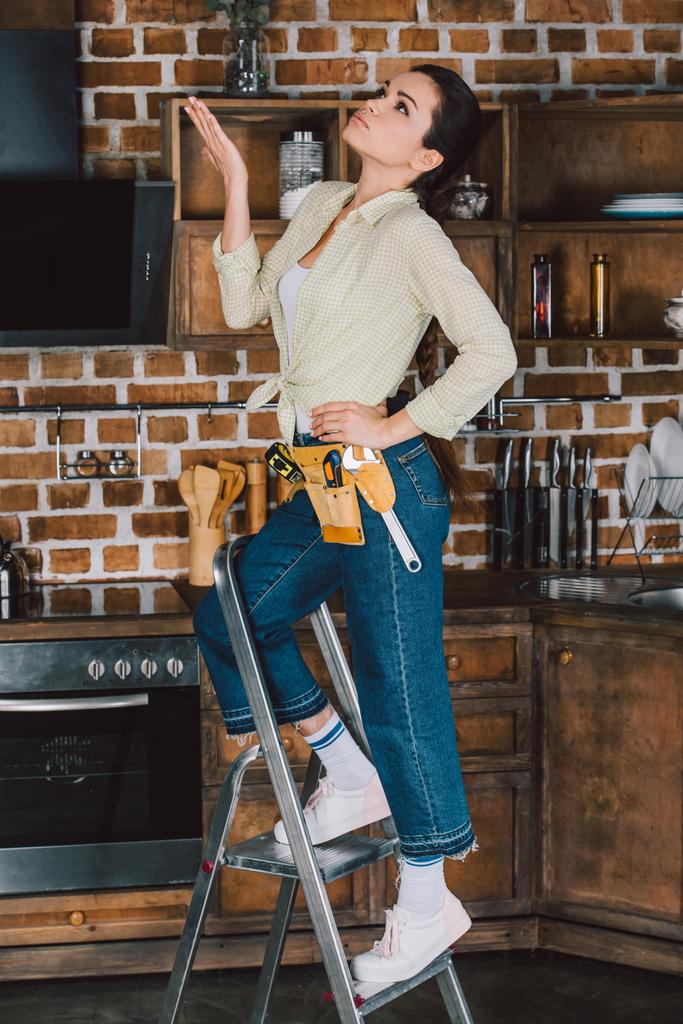 beautiful young repairwoman standing on stepladder at kitchen and looking up - Photo, Image