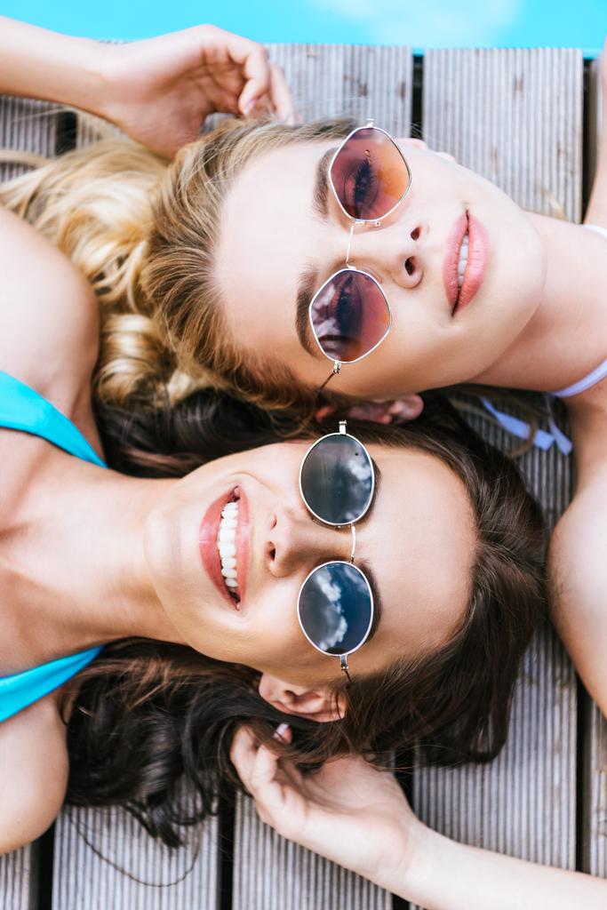 pohled shora krásných mladých žen v sluneční brýle se usmívá na kameru při ležení na bazénu  - Fotografie, Obrázek