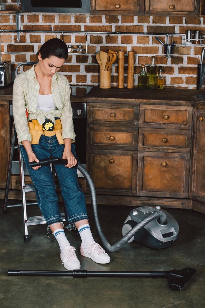 sad young repairwoman looking at broken vacuum cleaner while sitting on stepladder at kitchen - Photo, Image
