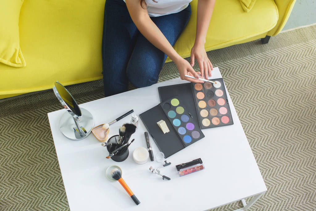 обрезанный снимок женщины с кистью макияжа сидя на диване за журнальным столиком с косметикой для макияжа дома
 - Фото, изображение