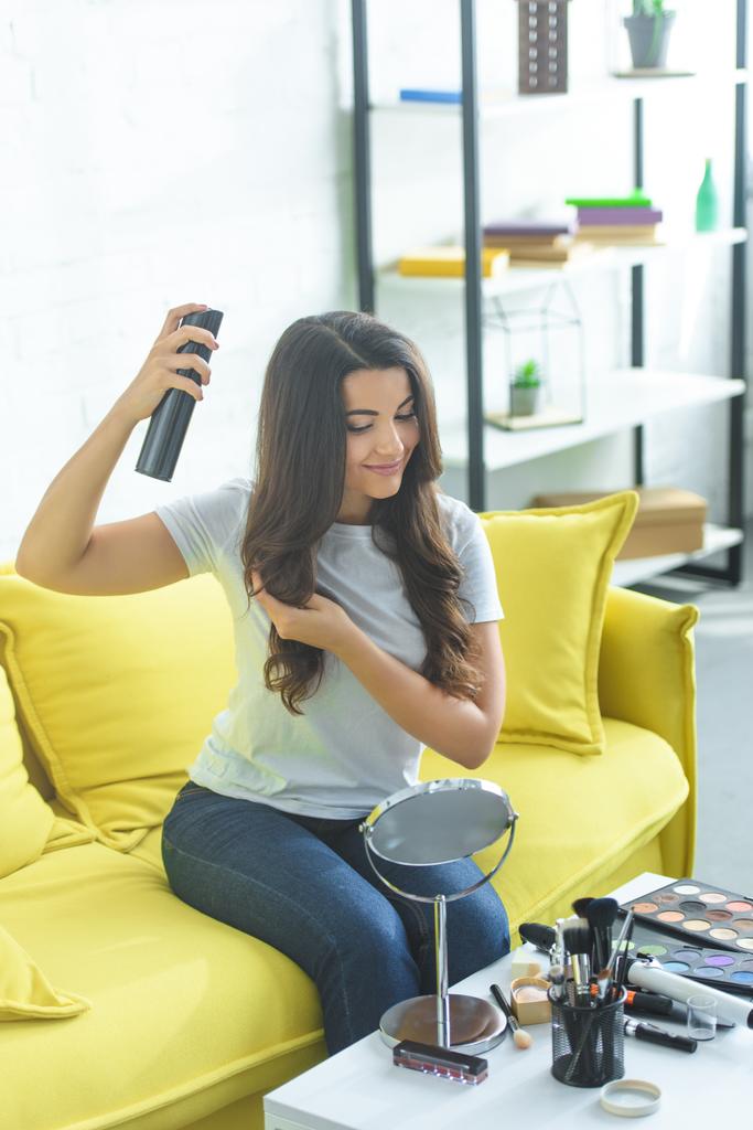 uśmiechnięta kobieta, stosując lakier do włosów, aby utrwalić fryzurę, siedząc na kanapie w domu - Zdjęcie, obraz