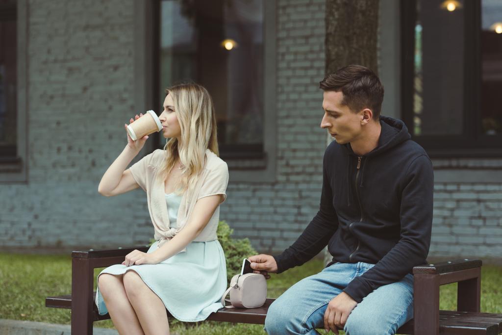 Mann klaut Smartphone aus Tasche einer Frau, die Kaffee trinkt, um auf Parkbank zu gehen   - Foto, Bild