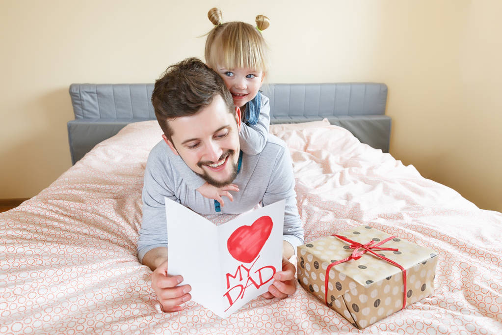 Vater und Tochter liegen an einem sonnigen Tag auf dem Bett. Sie überreicht ihrem Vater ein Geschenk und eine Grußkarte zum Vatertag. Selektiver Fokus.  - Foto, Bild